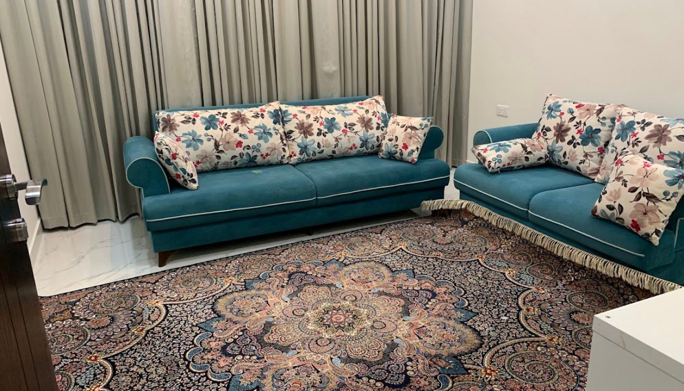 A living room with two teal sofas featuring floral cushions, a patterned rug, and beige curtains covering the window.