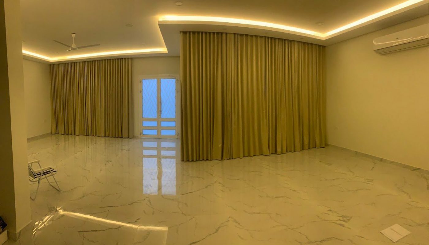 Spacious empty room with marble flooring, beige curtains covering windows and doors, ceiling lights, two ceiling fans, and a wall-mounted air conditioner.