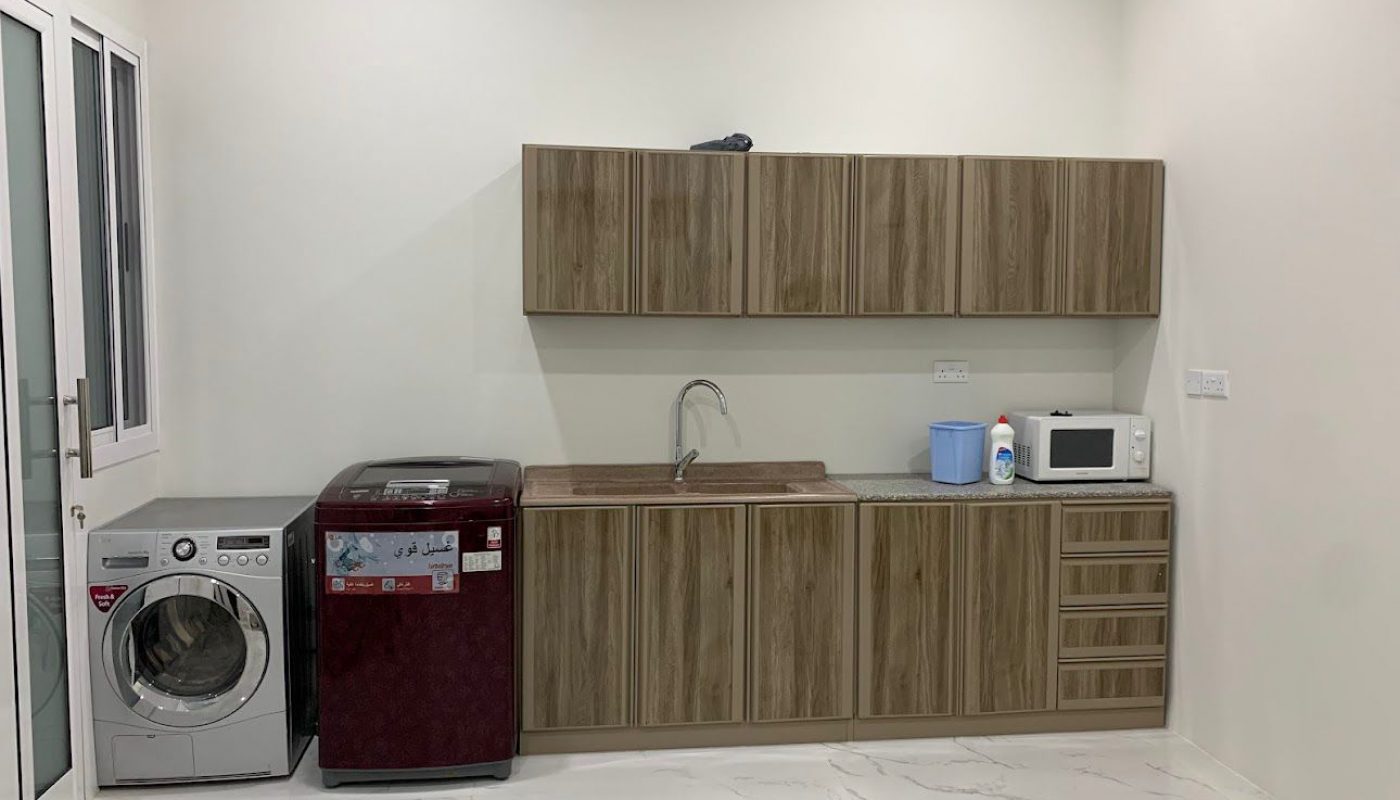A modern laundry room with wood cabinets, a sink, front-load washer, top-load washer, microwave, and cleaning supplies on a marble floor.