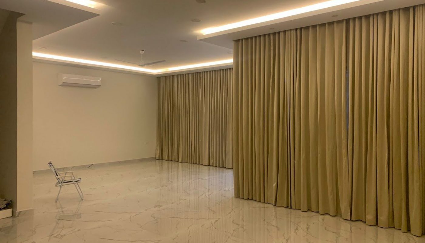 A spacious, empty room with marble flooring, beige curtains covering windows, ceiling fans, recessed lighting, and a single folding chair in the corner.