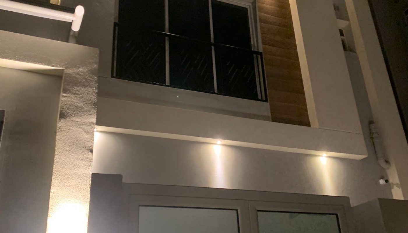 Modern two-story house exterior at night with illuminated lights, large windows, a metal and glass front door, and a wood accent panel beside the upper window.