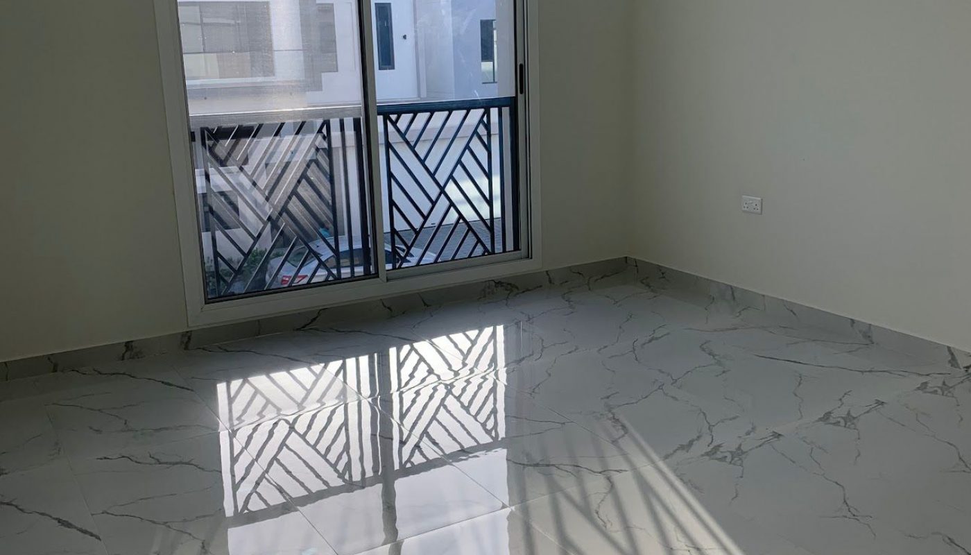Empty room with marble tile flooring, white walls, and a large sliding glass door leading to a balcony with patterned metal railing. Sunlight casts shadows on the floor.
