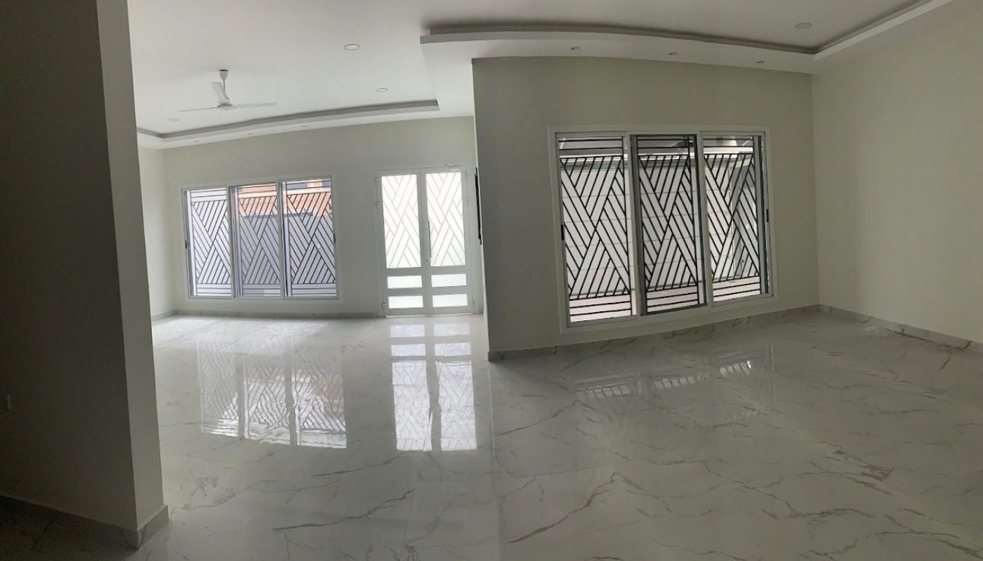 A spacious, empty room with white walls, marble floors, large windows, and glass doors with metal security grilles. Ceiling has recessed lighting and a ceiling fan.