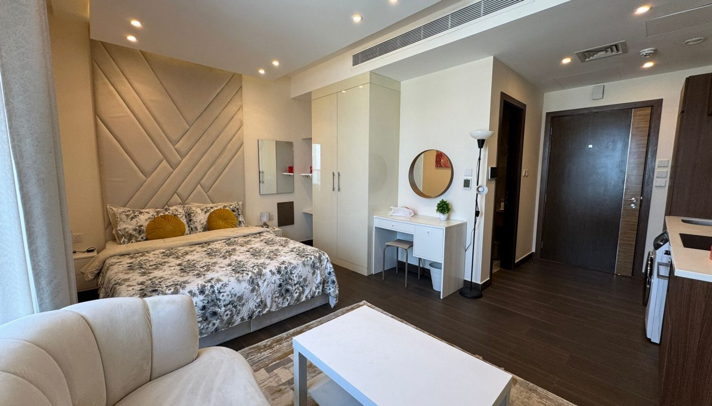 Modern studio apartment with a bed, small sofa, coffee table, desk, wardrobe, kitchenette, and wooden flooring. The room is well-lit with neutral tones and contemporary decor.