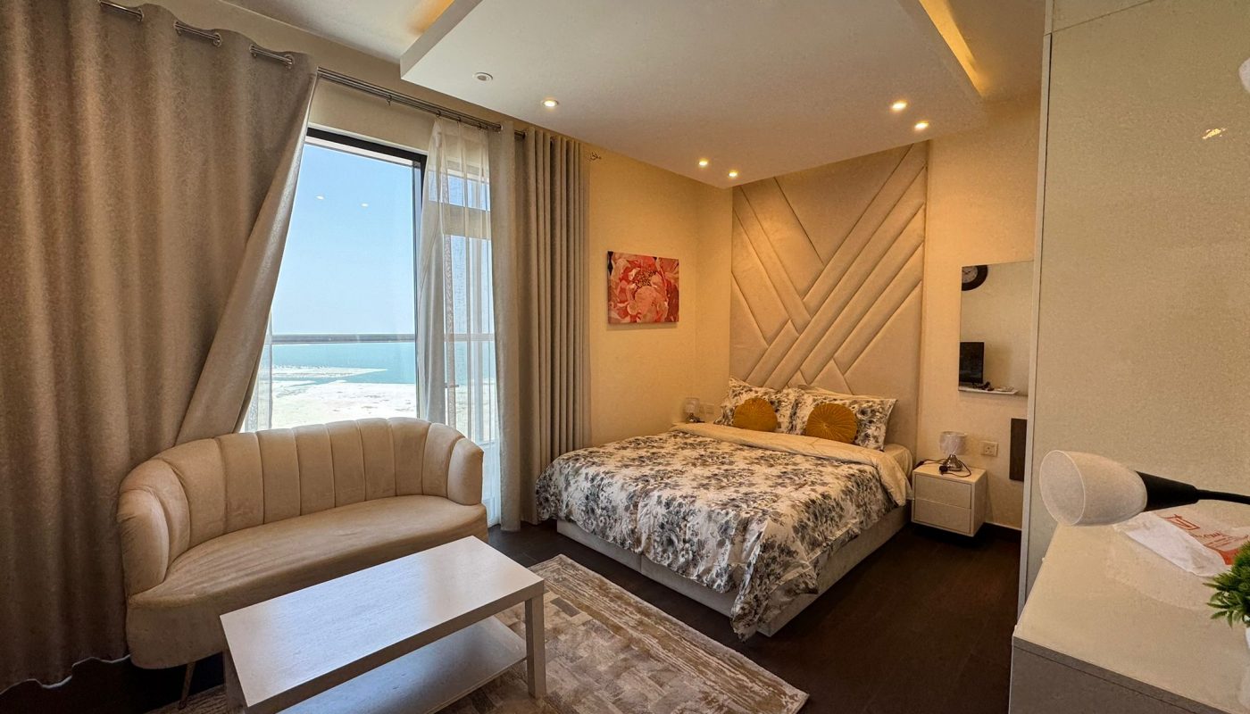 A modern bedroom with a large bed, patterned bedding, a cream sofa, a coffee table, and a window with a view of the sea. Neutral tones and soft lighting decorate the room.