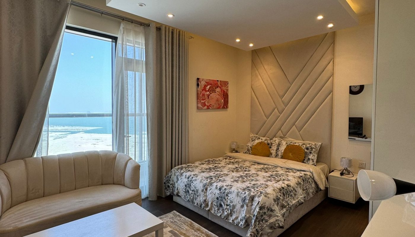 A modern bedroom with a large bed, patterned bedding, a white sofa, a coffee table, and floor-to-ceiling windows overlooking a bright outdoor scene.