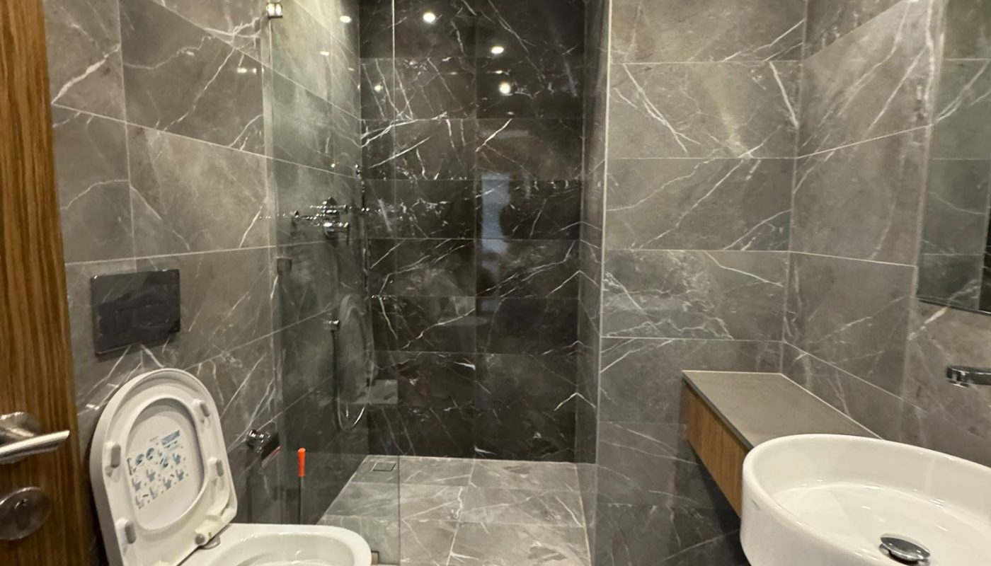 Modern bathroom with gray marble walls and floor, featuring a wall-mounted toilet with the lid up, a walk-in shower, a white sink, and wood accents.