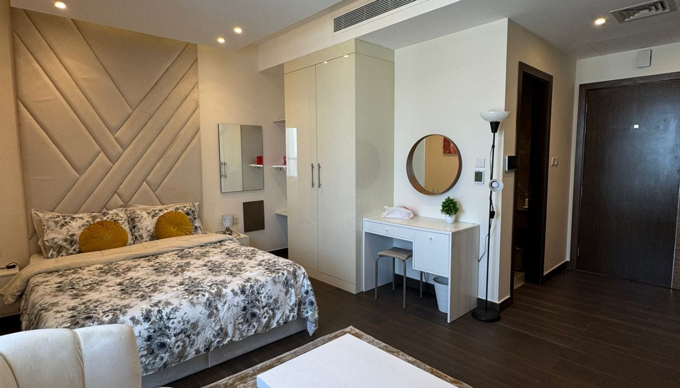 Modern hotel room with a large bed, white furniture, a round mirror on a desk, and a sofa, featuring neutral colors and contemporary decor.