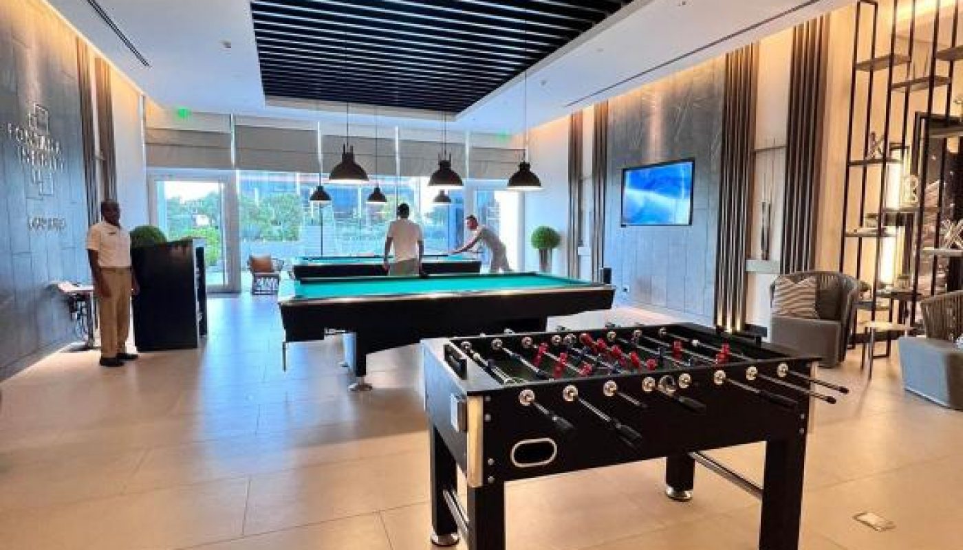 A modern game room with a foosball table in the foreground, a pool table in the background, and three people standing near large windows.