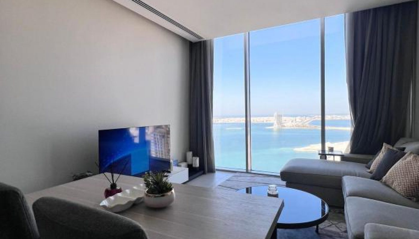 Modern living room with a dining table, sofa, TV, and large floor-to-ceiling windows overlooking a body of water and bridge in the distance.