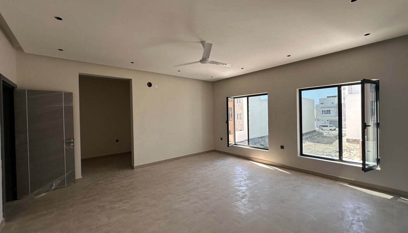 Spacious, empty room with tiled floor, ceiling fan, recessed lights, open windows, and neutral walls, receiving natural daylight from two large windows.