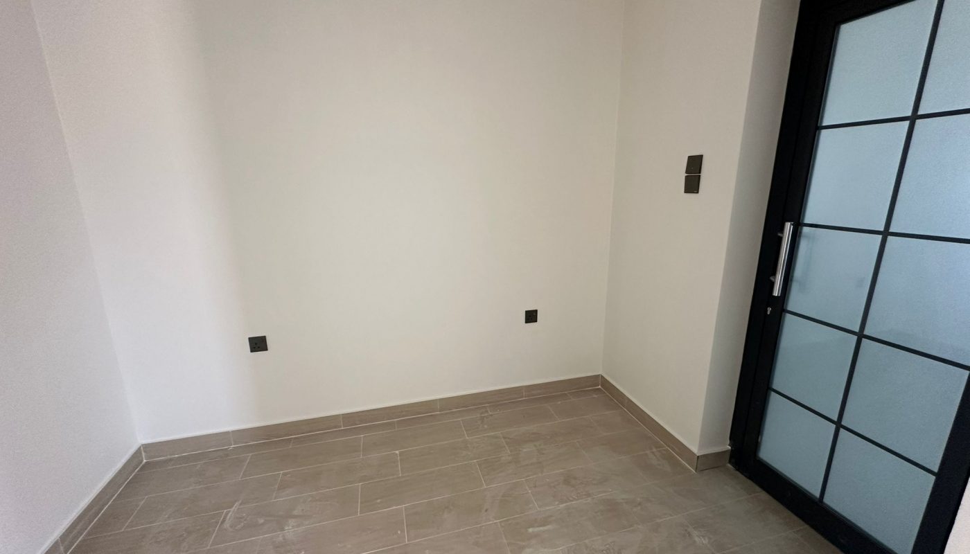 Small, empty room with light gray tiled floor, white walls, a black-framed glass door, and three visible electrical outlets.