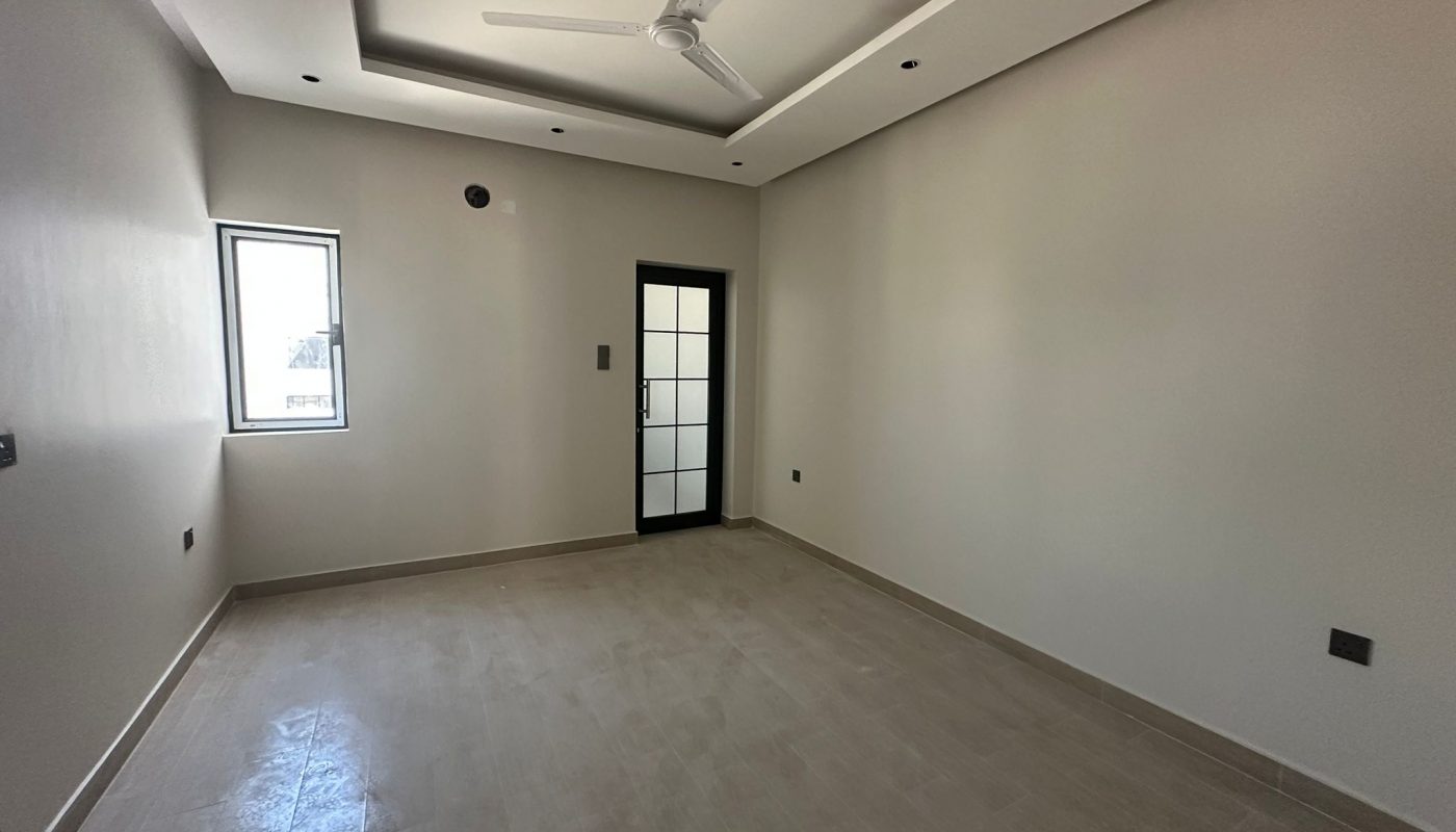 Empty room with beige tiled floor, off-white walls, a ceiling fan, a small window on the left wall, and a glass door at the back.