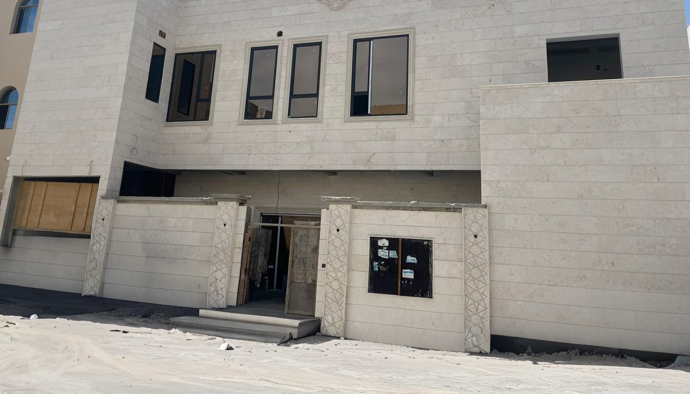 A modern, unfinished two-story building with large windows and a beige stone facade under a blue sky. Construction materials and sand are visible in the foreground.