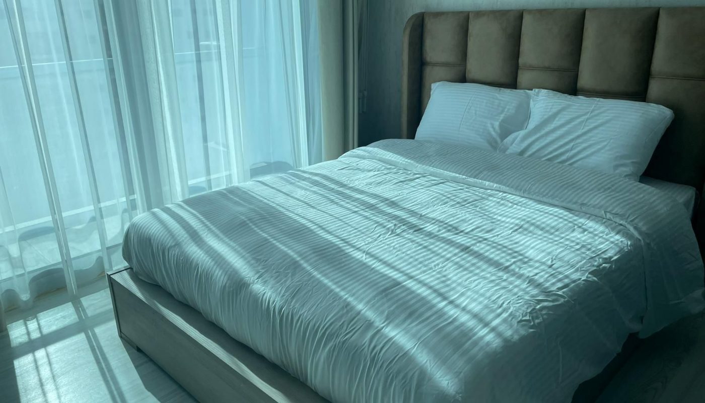 A neatly made bed with white linens is positioned near large windows covered by sheer curtains, allowing sunlight to cast shadows across the room.