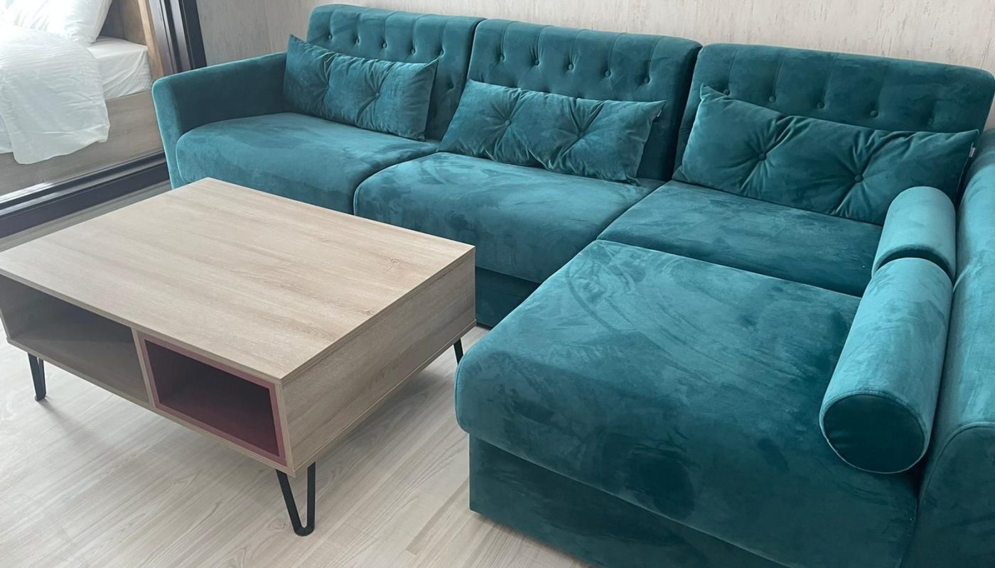 A teal velvet sectional sofa with tufted backrests and matching cushions is next to a light wood coffee table with open shelves on black metal legs. A bed is visible through a glass partition.