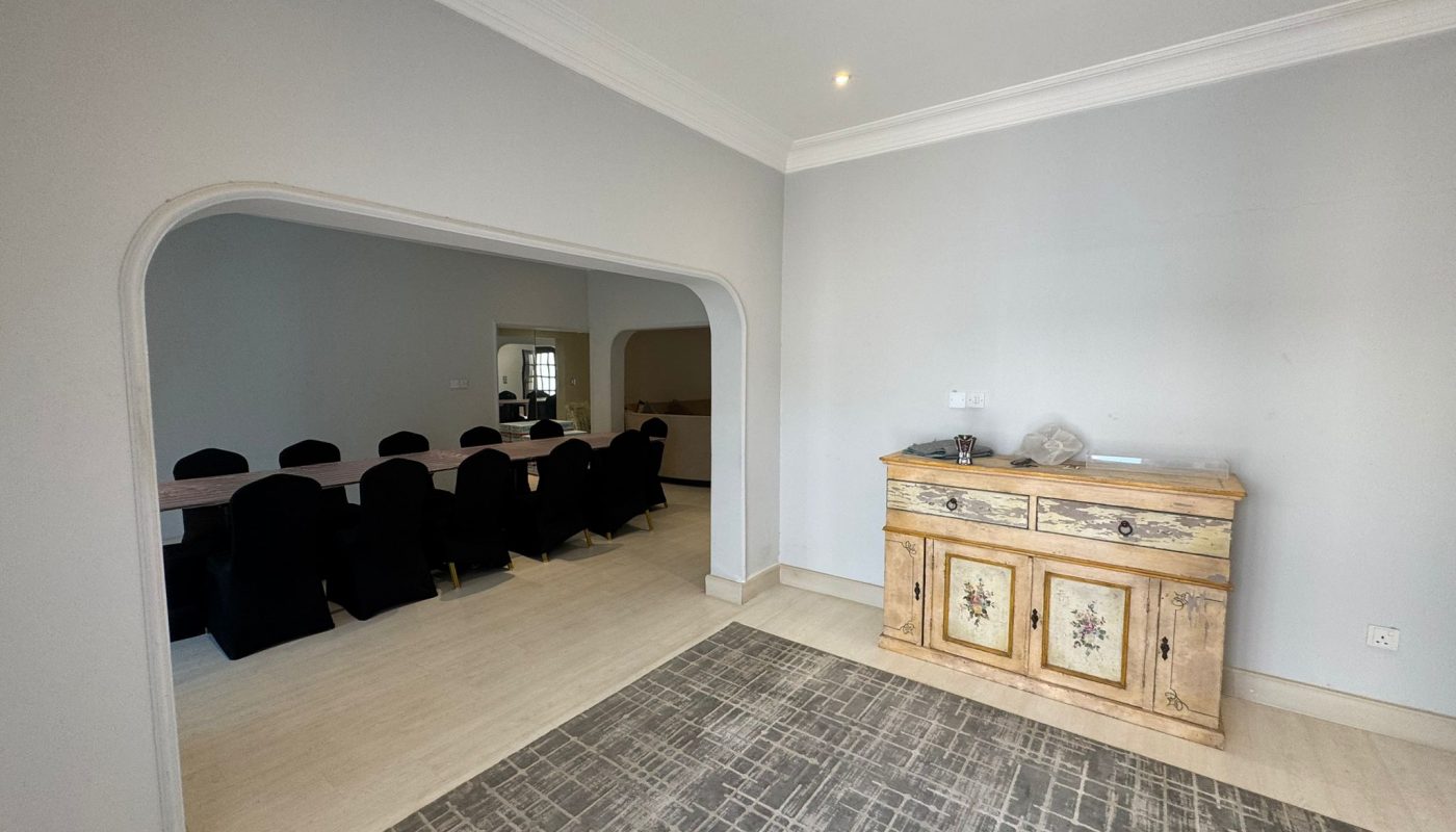 A room with a patterned rug, a wooden sideboard, and an open archway leading to a dining area with a long table and black-covered chairs.