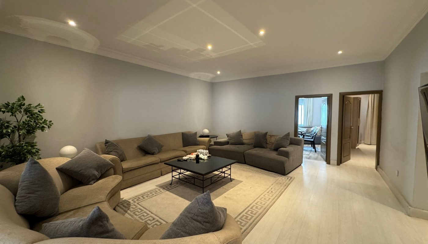 Modern living room with neutral-colored sofas arranged around a central coffee table, light wood flooring, minimal decor, and an open doorway leading to another room.