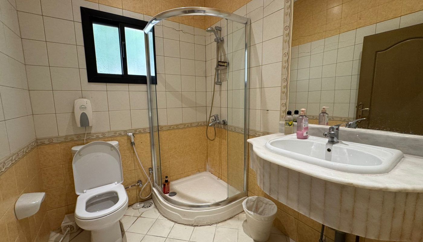 A bathroom with beige tiles, a glass shower enclosure, a toilet, a bidet, a sink with toiletries, a mirror, a waste bin, and a window.