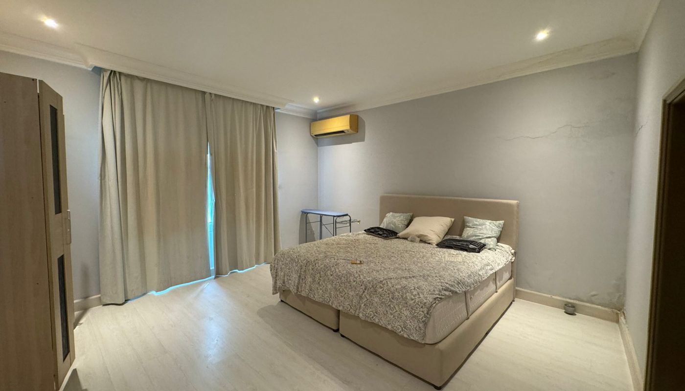 A simple bedroom with a double bed, two pillows, a wardrobe, a small desk, and closed beige curtains covering a window or door.