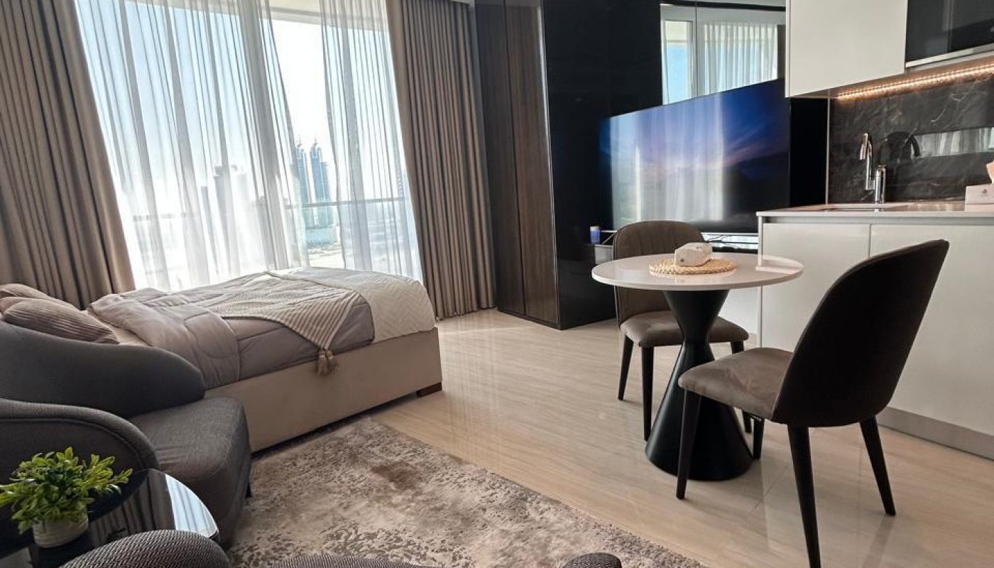 Modern studio apartment with a bed, sofa, dining table, and kitchen area. Large windows with sheer curtains let in natural light. Neutral tones and contemporary furnishings.