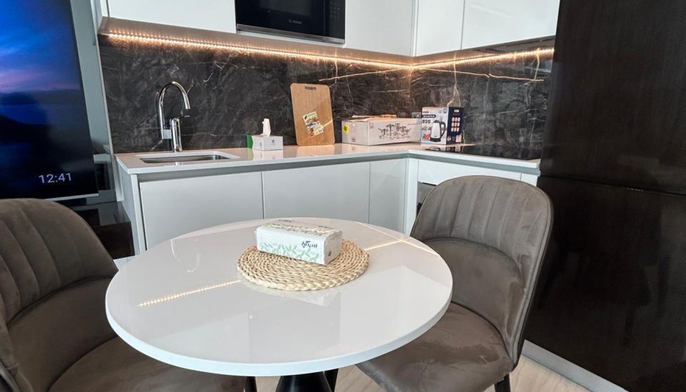 Modern kitchen with white cabinets, black marble backsplash, a round white table with two gray chairs, and tissue box on the table. Cardboard boxes are on the counter.