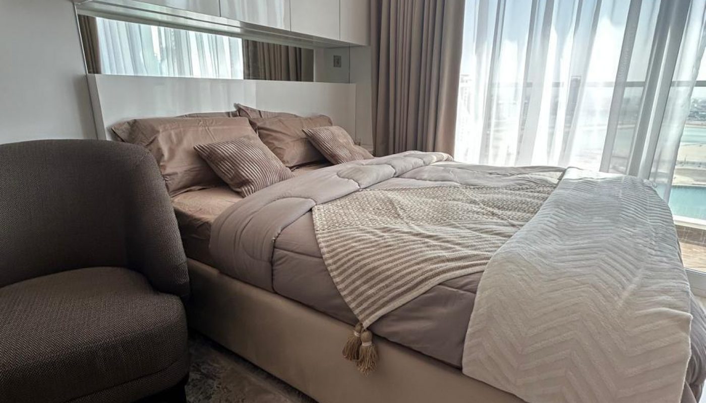 A neatly made bed with neutral bedding, a throw blanket, and several pillows next to a gray armchair in a bright bedroom with large windows and sheer curtains.