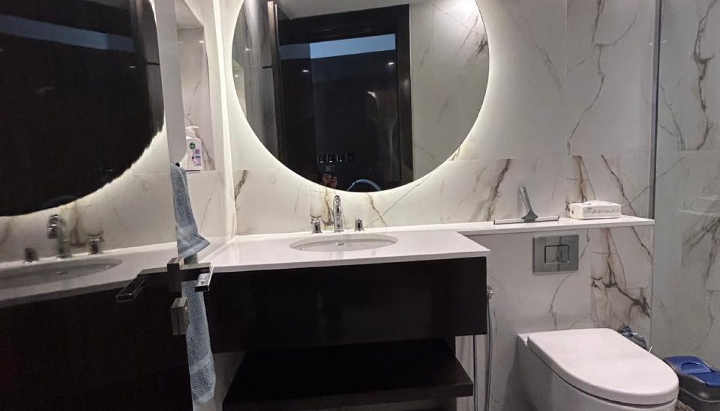 Modern bathroom with marble walls, a large round backlit mirror above a sink, wall-mounted toilet, and two black-and-white patterned rugs on the floor.