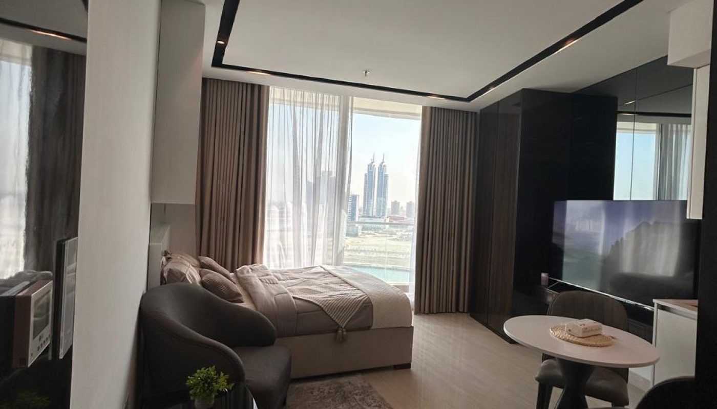 Modern studio apartment with a bed, two armchairs, a small dining table, TV, and large windows offering a city view.
