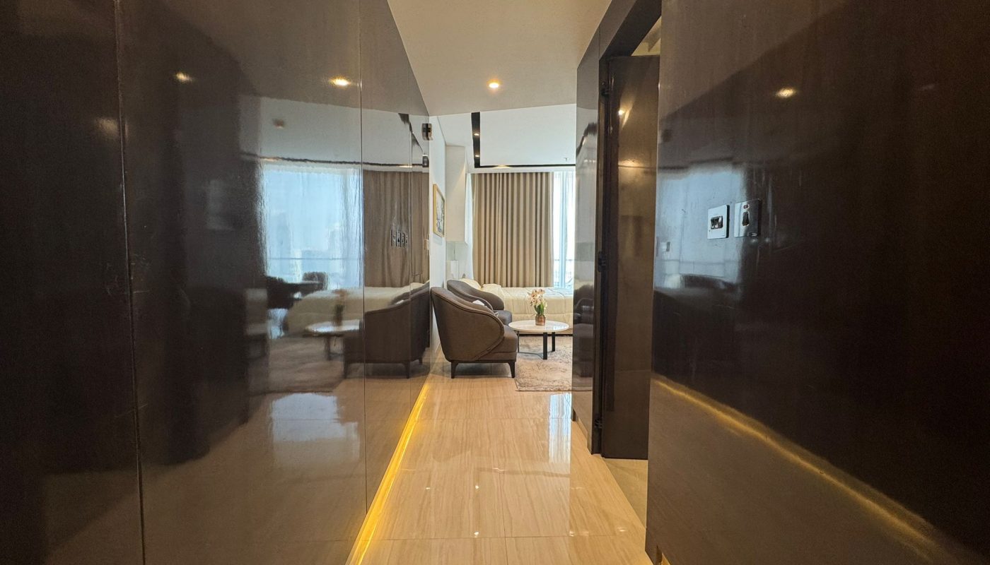 A polished hallway with dark walls and a light tiled floor leads to a modern living room with sofas, a coffee table, and large windows with curtains.