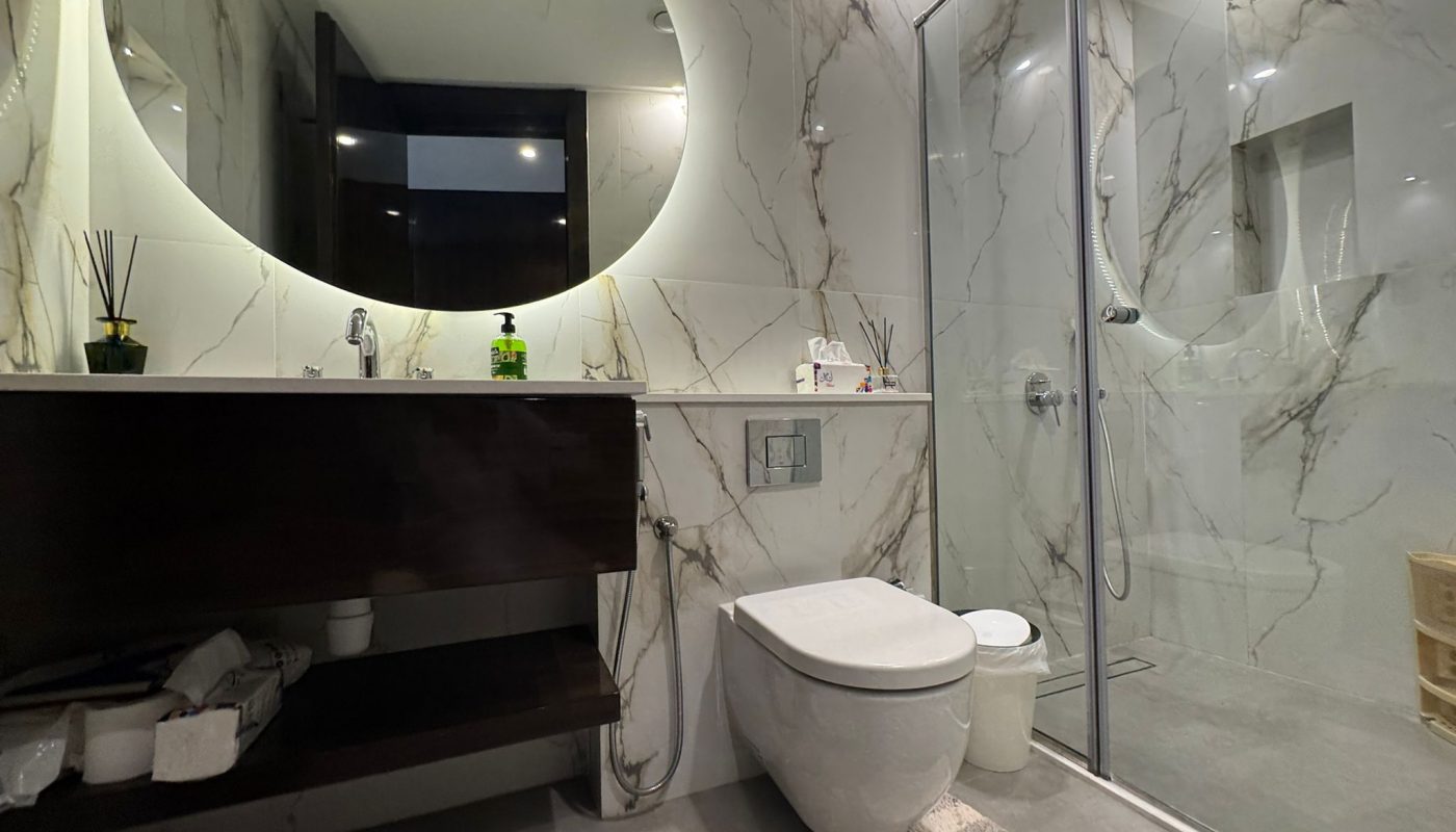 Modern bathroom with a large round backlit mirror, marble-patterned walls, a floating vanity, wall-mounted toilet, and glass-enclosed shower area.
