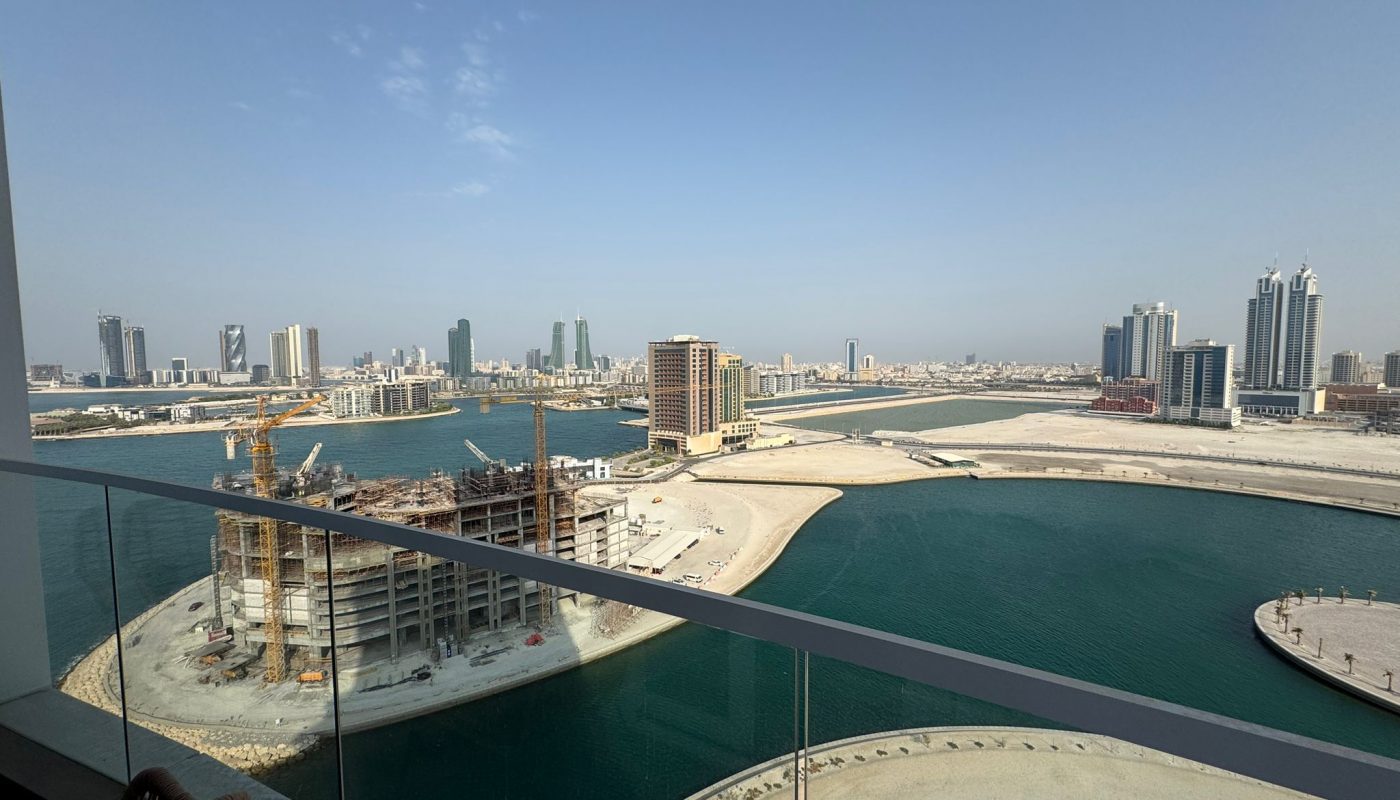 View from a balcony overlooking a waterfront with several under-construction and completed high-rise buildings in an urban cityscape under a clear blue sky.