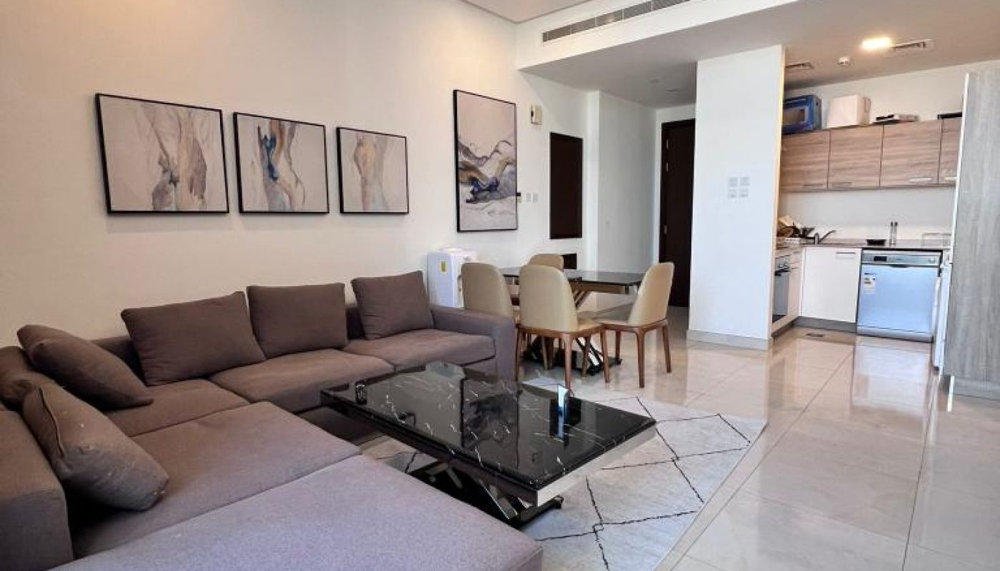 Modern open-plan living room and kitchen with a grey sectional sofa, glass coffee table, dining area, and neutral decor.