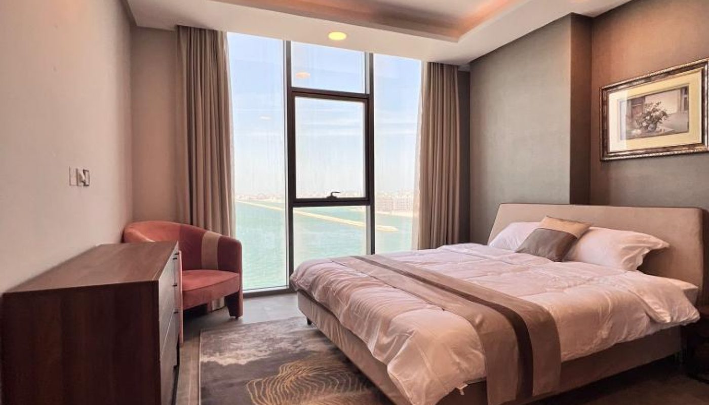 Modern bedroom with a large window overlooking water, a double bed with white linens, a wooden dresser, a pink chair, and a framed picture on a brown accent wall.