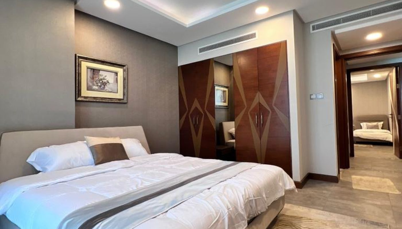 A modern bedroom with a large bed, wood-paneled wardrobe, gray walls, abstract rug, and framed artwork above the headboard. Another room is visible through an open doorway.
