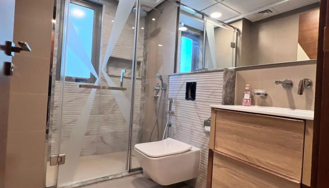Modern bathroom with a glass-enclosed shower, wall-mounted toilet, wooden vanity with sink, and large mirror. Walls and floor feature light-colored tiles.
