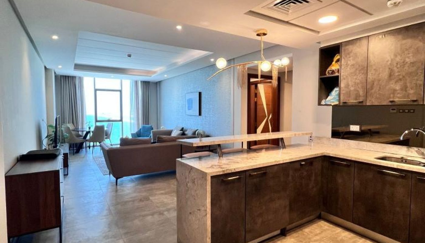 Modern open-plan kitchen and living room with tile flooring, brown cabinetry, a marble island, and large windows providing natural light.