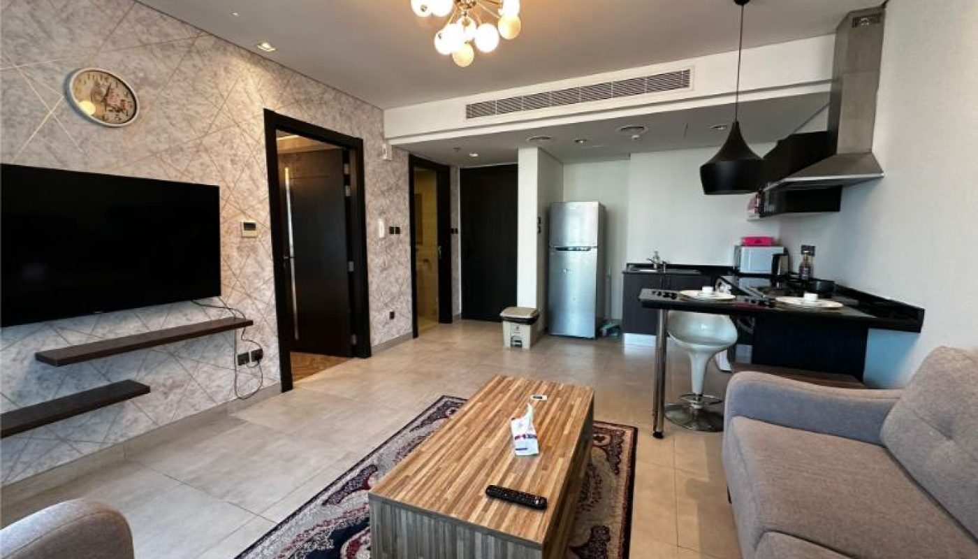 Modern living room and kitchen area with a wall-mounted TV, gray sofas, a wooden coffee table, and a small dining counter with two stools. A refrigerator and stove are visible in the kitchen.