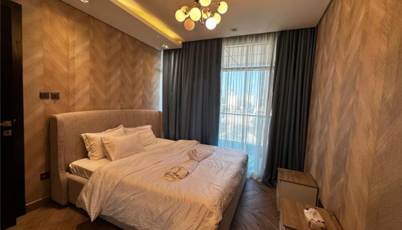 A modern bedroom with a double bed, beige headboard, white bedding, two nightstands, herringbone wood floors, and a window with sheer curtains and dark drapes.