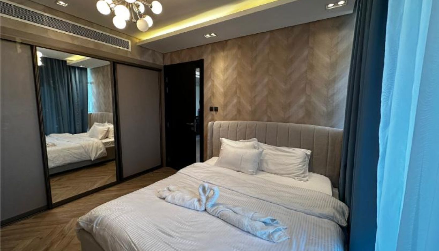Modern bedroom with a double bed, mirrored wardrobe, patterned accent wall, and a contemporary chandelier. Towels are folded on the neatly made bed.