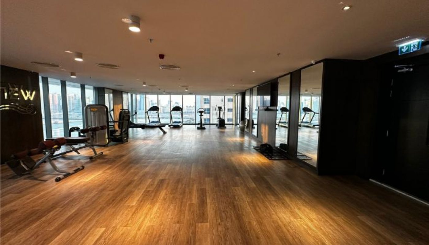 A spacious, well-lit gym with wood floors, fitness machines, and large windows overlooking a cityscape.
