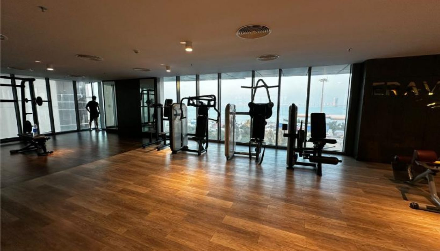 A modern gym with various exercise machines by large windows overlooking a cityscape; one person is standing near the windows on the left side.