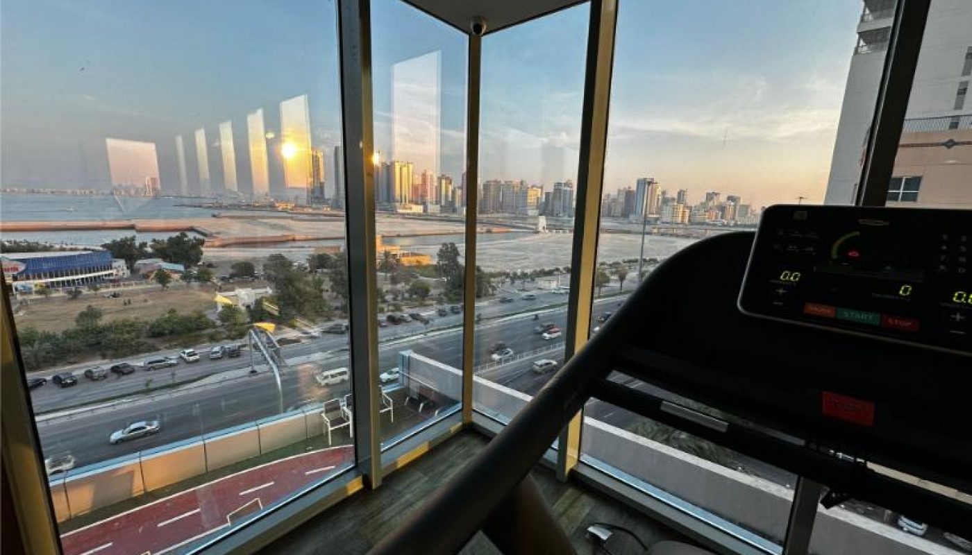 A treadmill is positioned near large windows overlooking a cityscape, busy road, and waterfront during sunset.