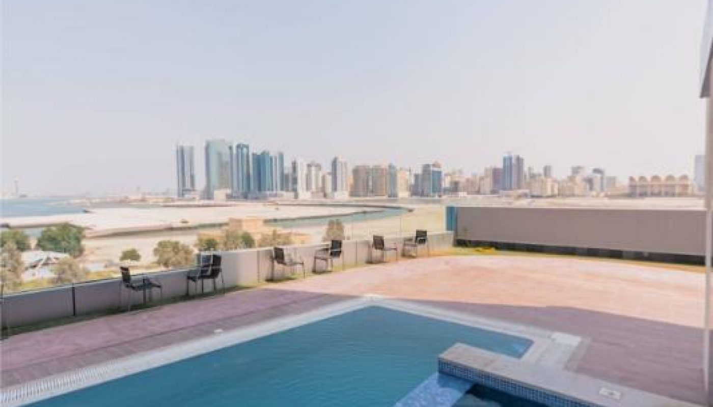 A rooftop pool with lounge chairs overlooks a city skyline with tall buildings and a body of water in the background under a clear sky.