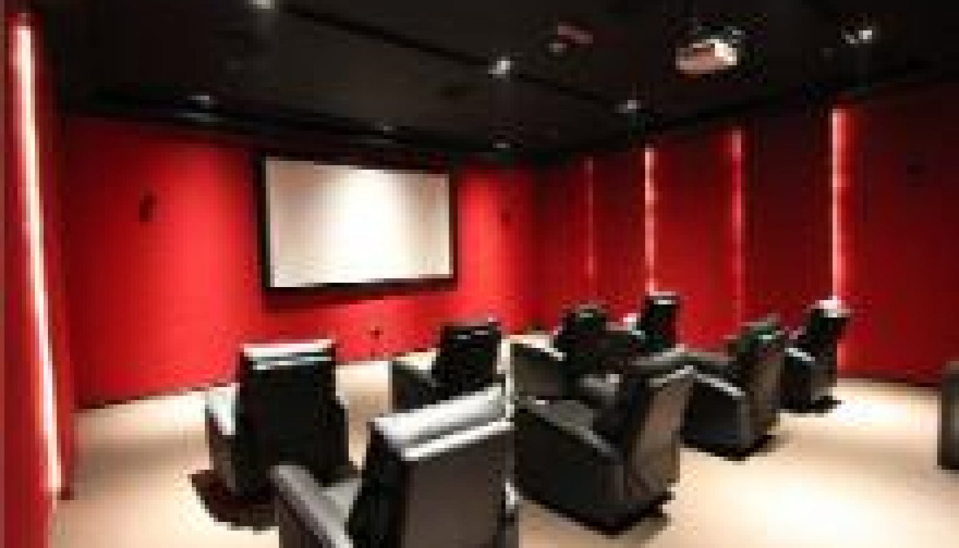 A small movie theater room with red walls, a large screen at the front, and several rows of black leather recliner chairs.