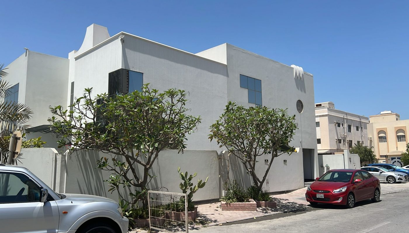 A modern white two-story house with large windows, surrounded by trees and a gray fence, sits next to parked cars on a sunny day.