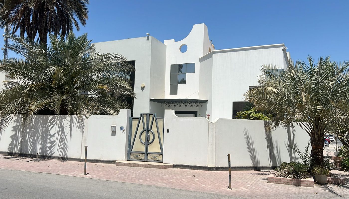 A modern white two-story house with geometric architectural features, surrounded by a white wall and metal gate, and bordered by palm trees under a clear blue sky.