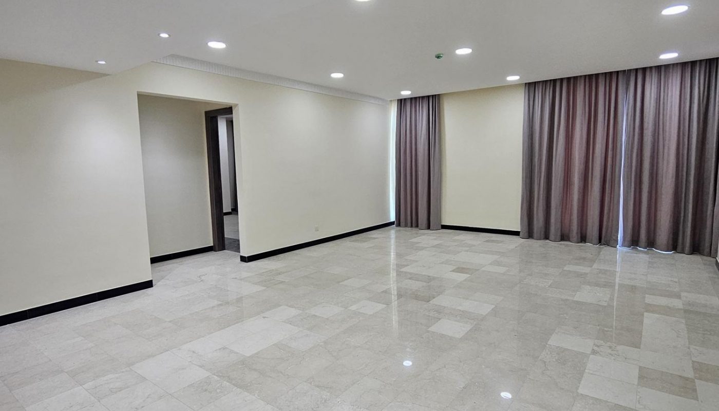 Spacious empty room with tiled floor, cream-colored walls, ceiling lights, and large windows covered by floor-length curtains.