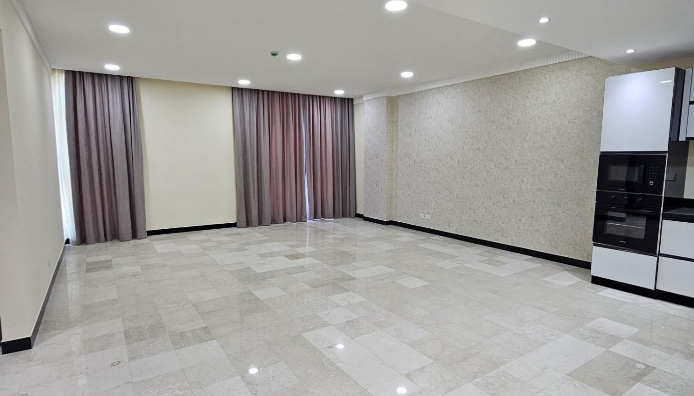 Spacious empty room with tiled floors, recessed ceiling lights, floor-to-ceiling windows covered by curtains, and a modern kitchen area on the right.