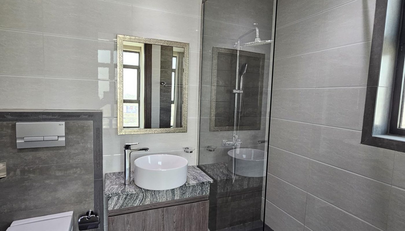 Modern bathroom with a wall-mounted toilet, a sink on a marble countertop, a large mirror, a walk-in shower with glass partition, and light gray tiled walls and floor.
